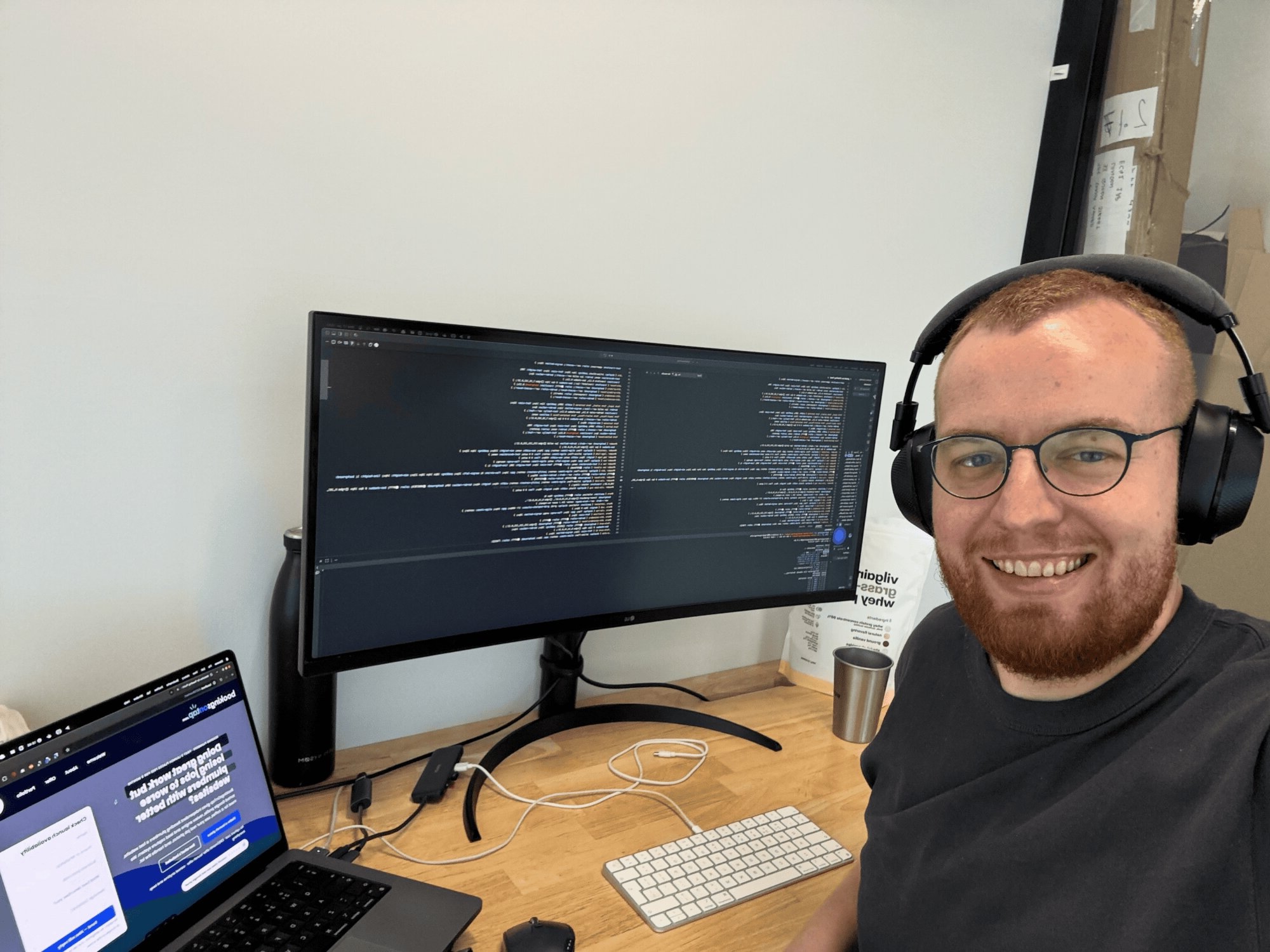 A man wearing headphones smiling at the camera, sitting in front of laptop and a large monitor with code on it
